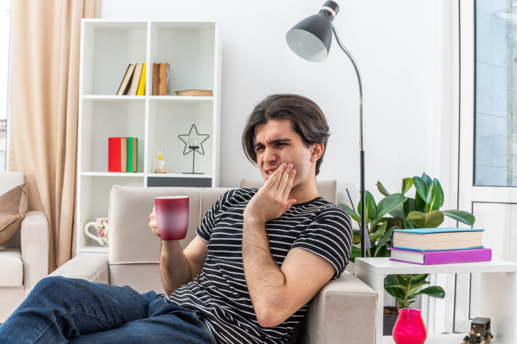 young-man-casual-clothes-holding-cup-hot-tea-looking-unwell-touching-his-cheek-feeling-toothache-sitting-chair-light-living-room